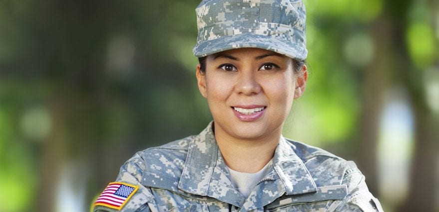 A young female member of the military dressed in camouflage jacket and hat is looking directly at the camera and smiling