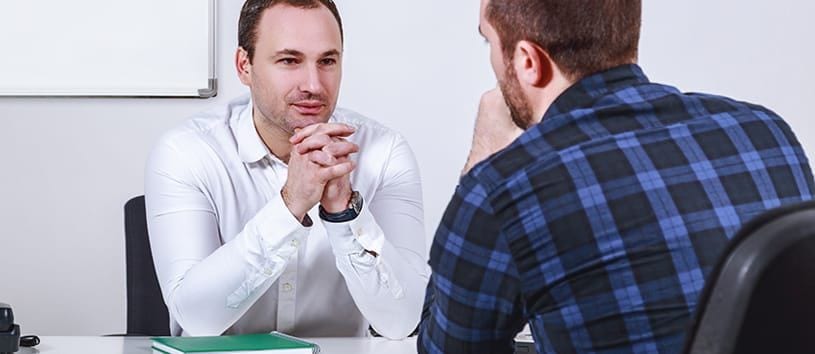 A man in a white collared shirt, presumably an admissions rep, sits across from another man with a trimmed beard, giving him his full attention